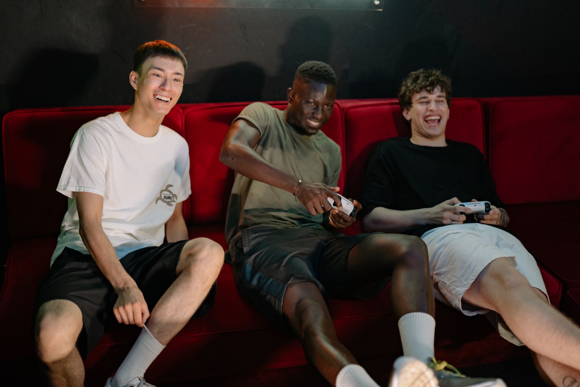 Three diverse young men sitting on a red couch, enjoying a fun video game session together. —